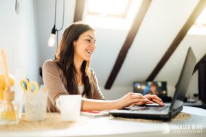 A woman smiles as she types on her laptop on a sunny day. This could symbolize treatment for adult ADHD in Columbus, OH. Contact Focused Mind ADHD Counseling for support with online therapy, an adult ADHD quiz, and other services!
