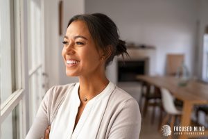 A woman smiles as she looks out the window. This represents overcoming ADHD and perfectionism in Columbus, OH. Contact a therapist at Focused Mind ADHD Counseling for support with questions like "what are anxiety symptoms" and more.