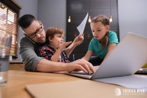 A parent struggles to type on their laptop while his children crowd around him. A therapist in Columbus Ohio can offer support for parents. Focused Mind ADHD Counseling can offer support for ADHD and emotional regulation and other services. Search ADHD therapist for adults near me to learn more!