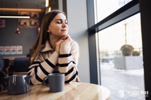 A woman smiles with her eyes closed next to a coffee shop window. Learn how Focused Mind ADHD Counseling can offer support in overcoming anxiety symptoms in Columbus, OH. We offer support for adhd and emotional regulation. Learn more about emotional regulation therapy in Columbus, OH today.
