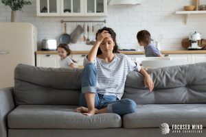 A mother holds her head as her children run in the kitchen behind her. Learn more about how ADHD and emotional regulation interact by searching "adhd specialist near me". We offer emotional regulation therapy in Columbus, OH and other services.