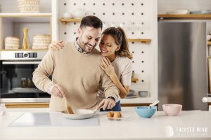 A couple smiles while cooking together in the kitchen. Learn how to address work related stress and learn more about ADHD and relationships today. Contact an adult ADHD therapist in Ohio to learn more about ADHD treatment for adults today.