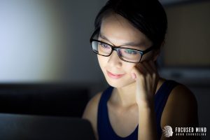 Young woman in glasses looking at her laptop in a dim room, researching ADHD testing for adults in Columbus, OH and connecting with an adult ADHD therapist in Ohio.