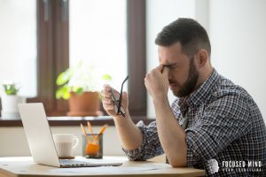 A man closes his eyes and pinches his nose bridge while holding his glasses, appearing stressed while working on a laptop. This scene is commonly seen by ADHD therapists in Columbus, OH when helping clients navigate ADHD and overthinking linked to RSD treatment in Columbus, OH.