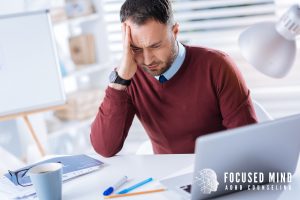 A man sits at his desk with his hand on his forehead, appearing overwhelmed while working on his laptop. This reflects common struggles with ADHD and emotional regulation, especially when pressure builds. He may be considering emotional regulation therapy in Columbus, OH or support from a Columbus ADHD therapist who understands adult ADHD challenges.