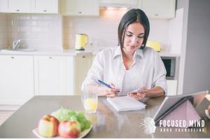 A woman sits at her kitchen table writing in a notebook while referencing her tablet, demonstrating an organized strategy to manage daily tasks—often essential for adults with ADHD. This scene reflects skills learned through adult ADHD treatment in Columbus, OH or with support from an adult ADHD specialist in Ohio. It also shows how improving routines can support better ADHD and emotional regulation outcomes.