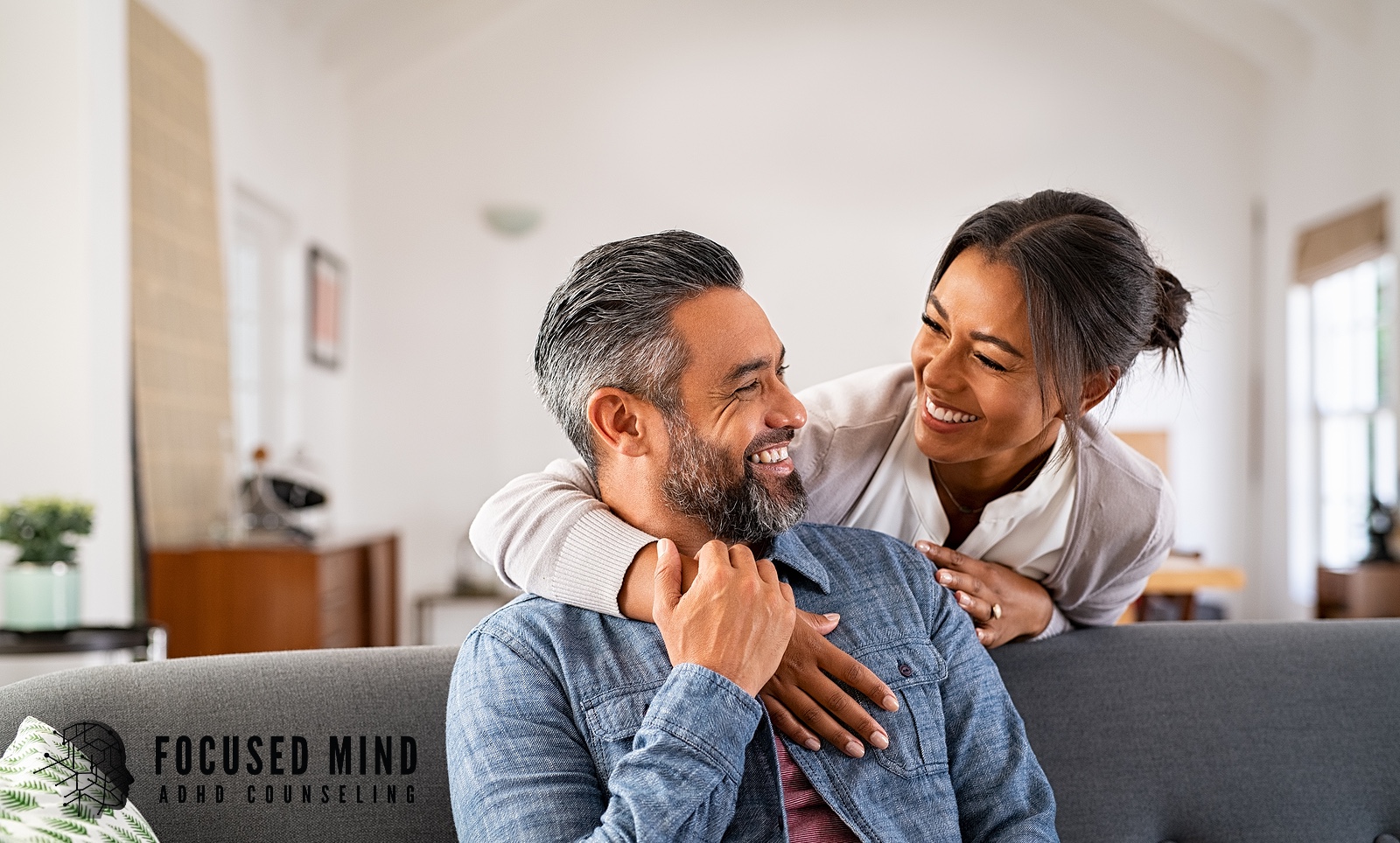 A woman embraces her smiling partner from behind on a couch. Can understanding ADHD's role in your marriage restore the closeness you've been missing? Online ADHD testing in Cincinnati, OH, offers answers that help couples move from frustration to empathy and reconnection.