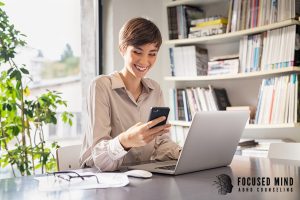 Woman smiling while checking her phone at a laptop, representing relief that can come with the right support. Online adhd treatment in columbus, oh, an adhd therapist in columbus, oh, and adult adhd treatment in columbus, oh can improve follow-through and balance.