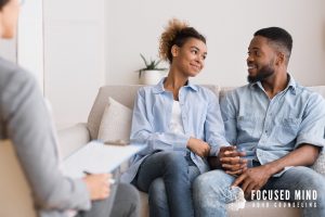A couple sits closely together on a couch, smiling warmly at each other during a therapy session, with a therapist taking notes nearby. This sense of ease and connection reflects what ADHD treatment for adults in Columbus, Ohio helps couples build over time — a relationship where differences are understood rather than resented. For those searching for an ADHD therapist for adults near me, this kind of supportive, ADHD-informed environment is exactly what Focused Mind ADHD Counseling provides.