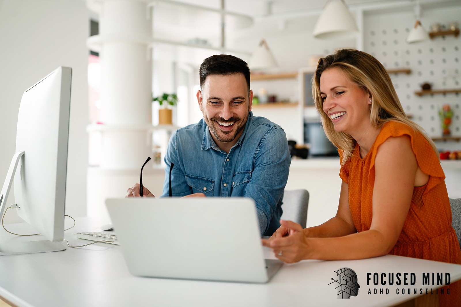 A man and woman sit together at a desk, smiling and laughing while looking at a laptop screen in a bright, modern space. This kind of genuine connection is what ADHD therapy in Columbus, OH helps couples work toward — moving past the frustration and finding their way back to the partnership that brought them together. An ADHD counselor in Columbus, OH can help couples build the systems and communication tools that make moments like this one more consistent.