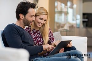 A couple sits closely together on a couch, smiling and engaged as they look at a tablet screen, suggesting shared planning or a relaxed moment of connection at home. Using shared digital tools like calendars and task lists is one of the practical strategies recommended in online ADHD treatment in Columbus, OH for couples navigating ADHD together. Adult ADHD treatment in Ohio helps partners build systems like these that work with the ADHD brain rather than against it.