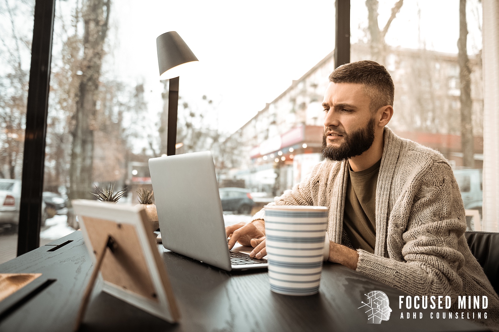 A bearded man works intently on a laptop at a café table beside a striped mug and warm lamp light. Is fear of failure showing up as avoidance, procrastination, or an inability to start tasks that matter to you? An ADHD specialist in Cincinnati, OH can help you get to the root of it.