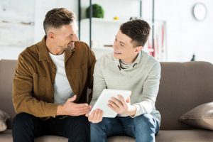 A father and teenage son sit together on the couch smiling while looking at a tablet, the kind of positive connection that can grow once a family understands adhd testing for teens in Columbus, OH results. RSD treatment in Columbus, OH or an online adhd therapist Cincinnati can support this progress
